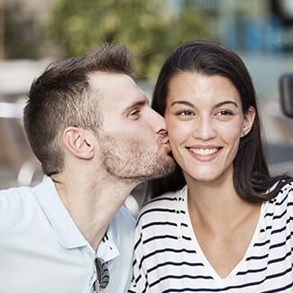 Woman with brown hair smiling as she gets kissed by her boyfriend on the cheek.