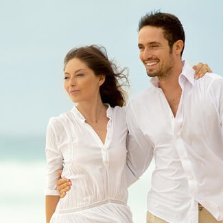 A man and woman wearing white clothes and hands around each other as they walk on the beach.
