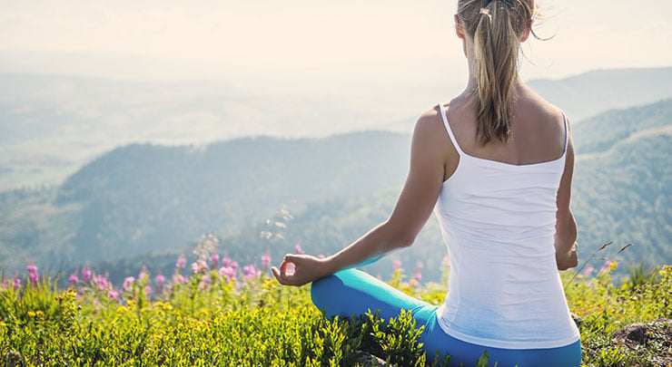 Woman in white tank top and blue pants sitting on grassy mountain meditating after clitoral hood reduction.